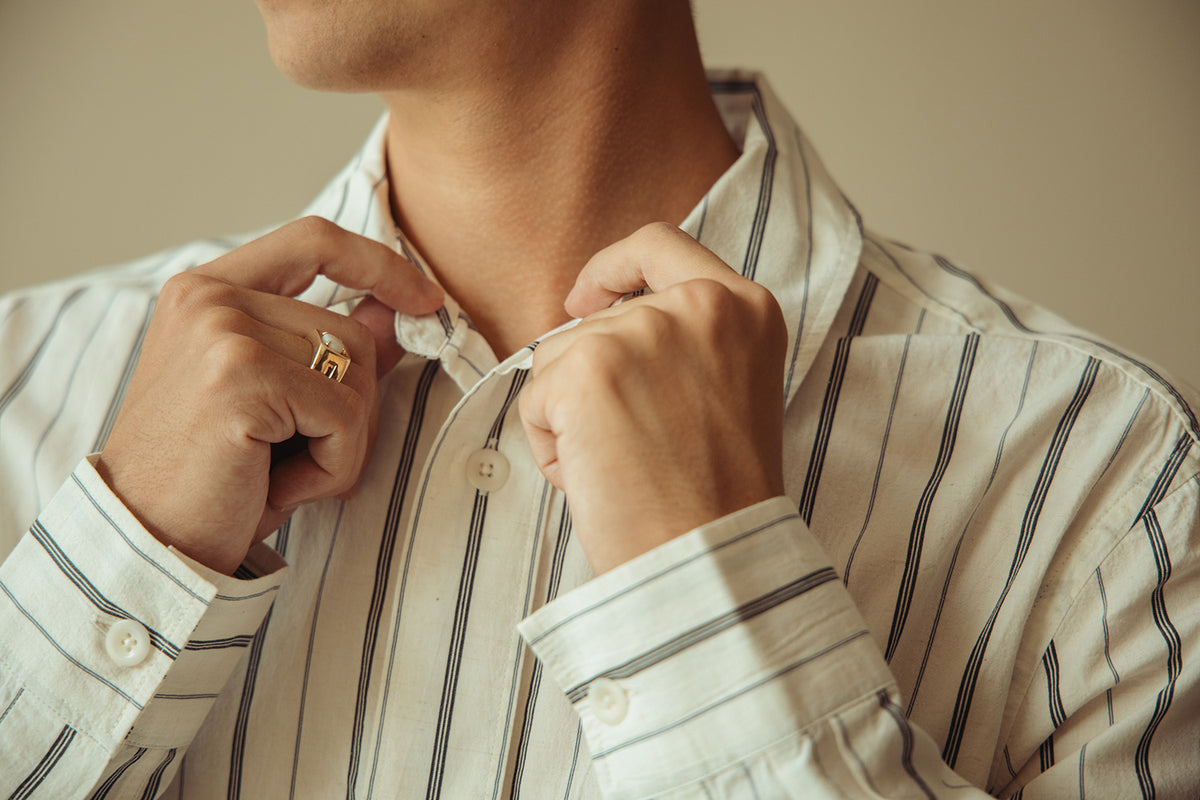Close-up of a man adjusting the collar of a white button-down shirt with thin vertical navy stripes. He is wearing a gold ring on one finger, and the background is a soft, warm beige.