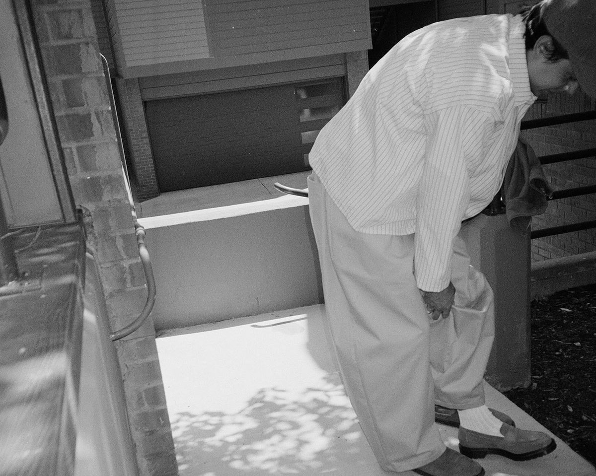 A black and white photograph of a man in a light-colored striped shirt and trousers bending forward on a porch step.